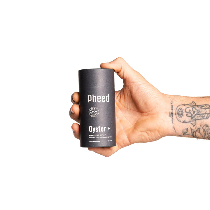 Tattooed hand holding a Pheed Oyster+ capsule bottle against a white background.