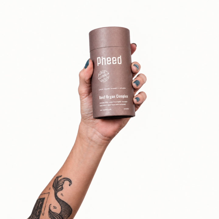 Tattooed hand holding Pheed Beef Organ Complex capsules on a white background.