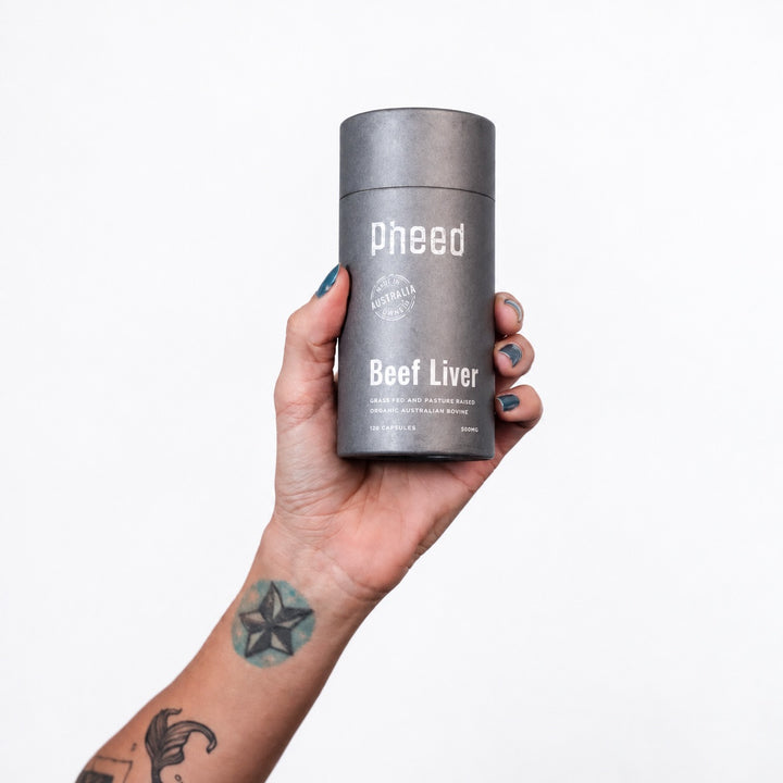 Tattooed hand holding a Pheed Beef Liver Capsules bottle on a white background.