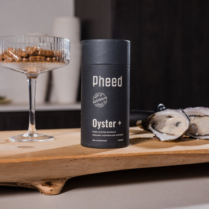 Pheed Oyster+ capsule bottle beside a glass filled with capsules and fresh oysters on a wooden board.