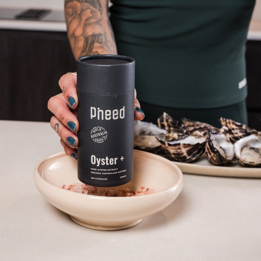 Woman holding a Pheed Oyster+ capsule bottle above a bowl of pink salt, with fresh oysters behind it.