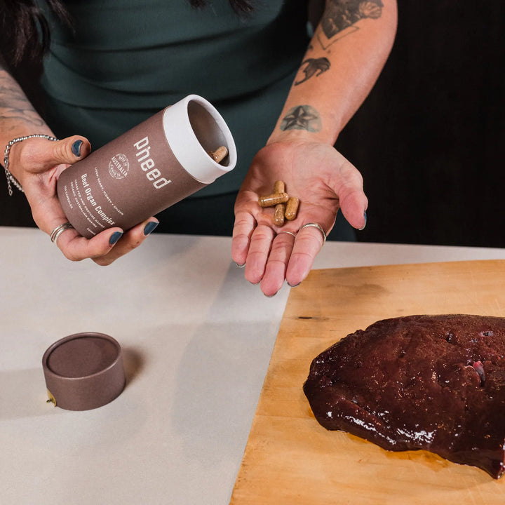 Pheed Beef Organ Complex capsules in an open palm beside the tube and raw beef liver on a chopping board.